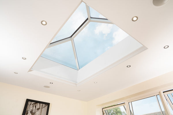 Modern room ceiling with a glass roof lantern showing blue sky and clouds, surrounded by recessed ceiling lights.